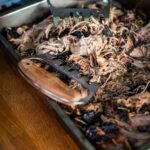 Zartes Pulled Pork aus dem Smoker, serviert auf einem Holzbrett mit BBQ-Soße und Burgerbrötchen.