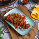 Schaschlik Spieße mit saftigem Schweinefleisch, Paprika und Zwiebeln, serviert in einer würzigen Tomatensoße. Im Hintergrund ein Teller mit dampfendem Reis – ein herzhaftes Schaschlik Rezept, perfekt für ein gemütliches Essen.