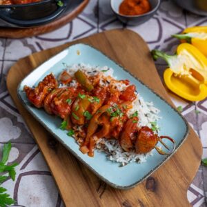 Schaschlik Spieße mit saftigem Schweinefleisch, Paprika und Zwiebeln, serviert in einer würzigen Tomatensoße. Im Hintergrund ein Teller mit dampfendem Reis – ein herzhaftes Schaschlik Rezept, perfekt für ein gemütliches Essen.