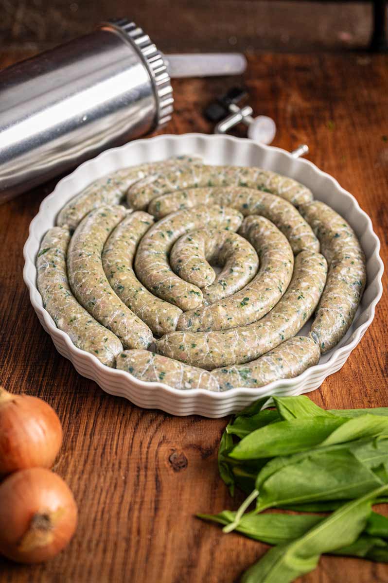 Roh gefüllte Bärlauch Bratwürste auf einem Holzbrett, umgeben von frischem Bärlauch und Gewürzen, bereit für den Grill oder die Pfanne.