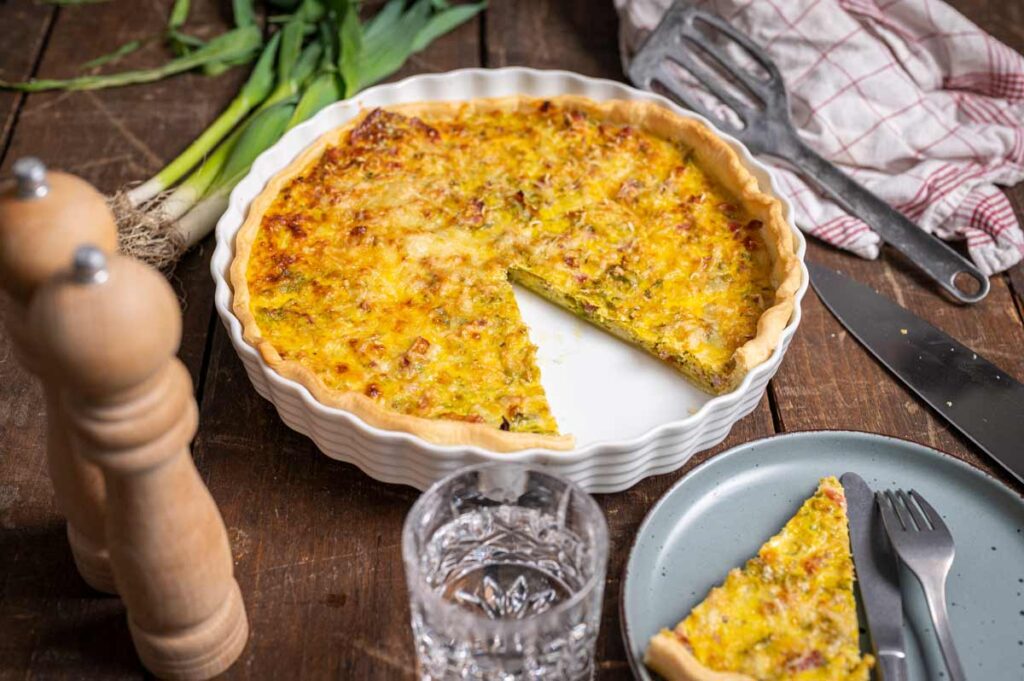 Eine frisch gebackene Lauch Quiche mit goldbrauner Käsekruste in einer Tarteform, serviert mit einem Messer auf einem Holzbrett.