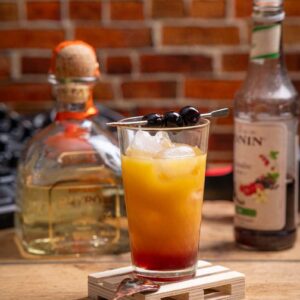 Ein leuchtender Tequila Sunrise Cocktail mit Tequila, Orangensaft und Grenadine, serviert auf Eis und garniert mit einer Maraschino Kirsche und Orangenzeste.