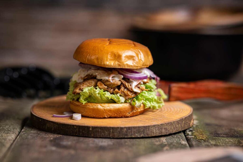 Nahaufnahme eines hausgemachten Pulled Chicken Burgers, angerichtet auf einem rustikalen Holzbrett mit BBQ-Sauce und knackigem Cole Slaw