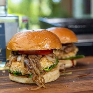 Vegetarischer Burger mit paniertem Grillkäse und Whisky-Zwiebel-Champignon-Mix, angerichtet auf einem Holzbrett.