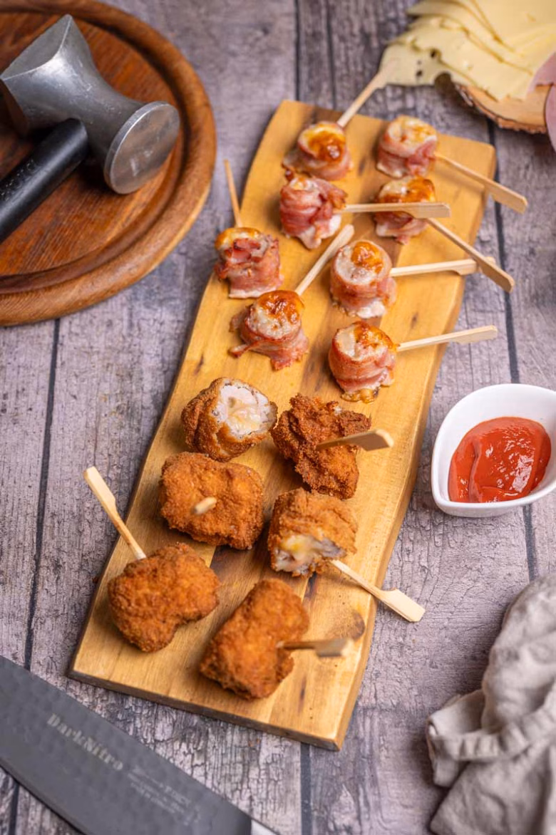 Zwei Varianten von Cordon Bleu Spießen auf einem Holzbrett, einmal mit knusprigem Bacon, einmal paniert und frittiert