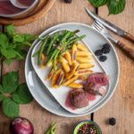 Zart rosa Rehrücken mit fruchtiger Brombeersoße, grünen Bohnen und knusprigen Schupfnudeln auf einem Teller