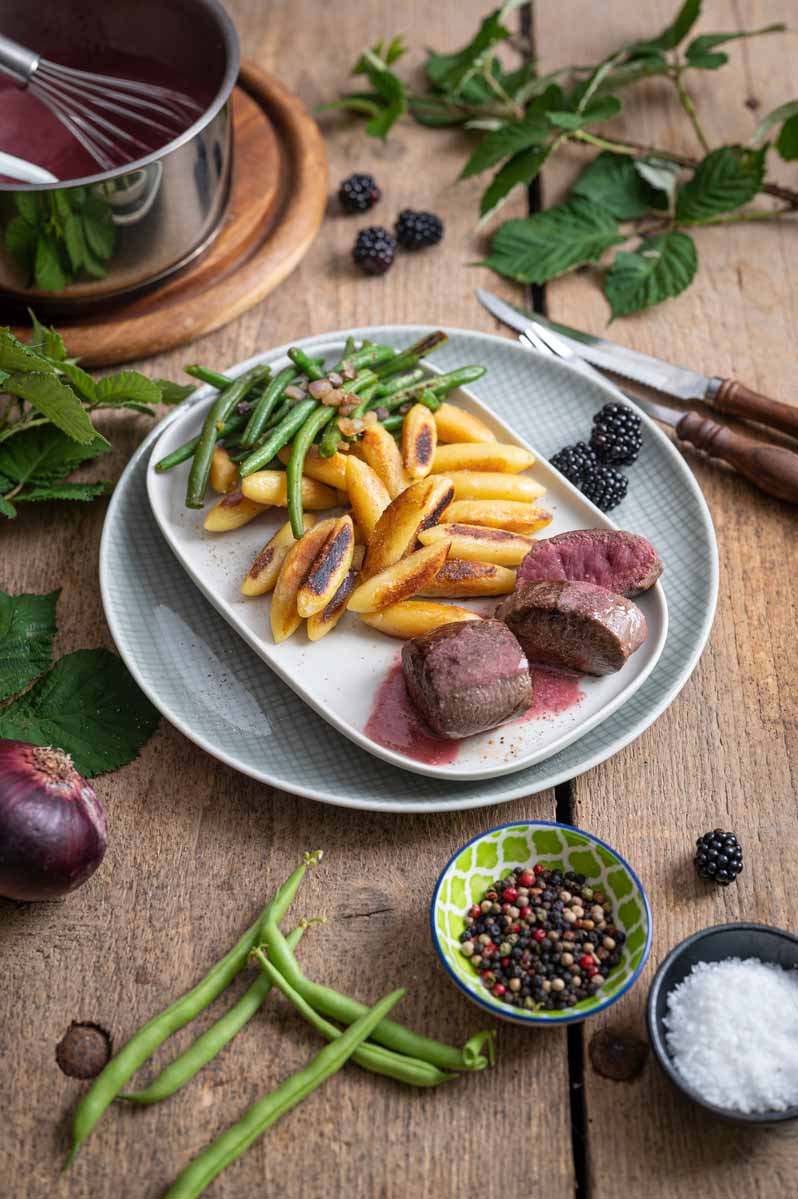 Zart rosa Rehrücken mit fruchtiger Brombeersoße, grünen Bohnen und knusprigen Schupfnudeln auf einem Teller