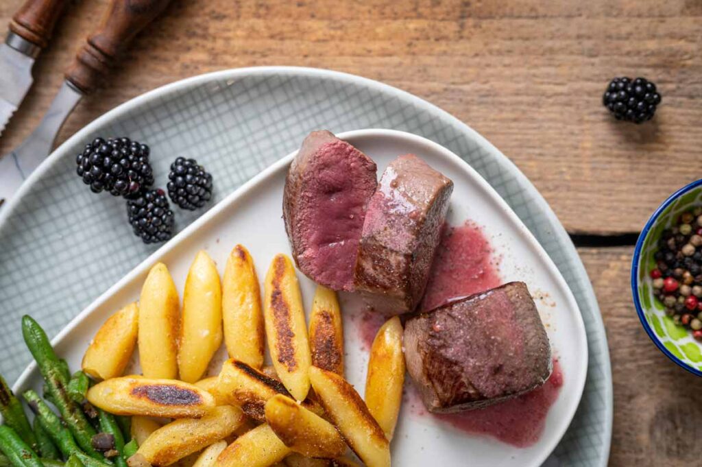 Zart rosa Rehrücken mit fruchtiger Brombeersoße, grünen Bohnen und knusprigen Schupfnudeln auf einem Teller