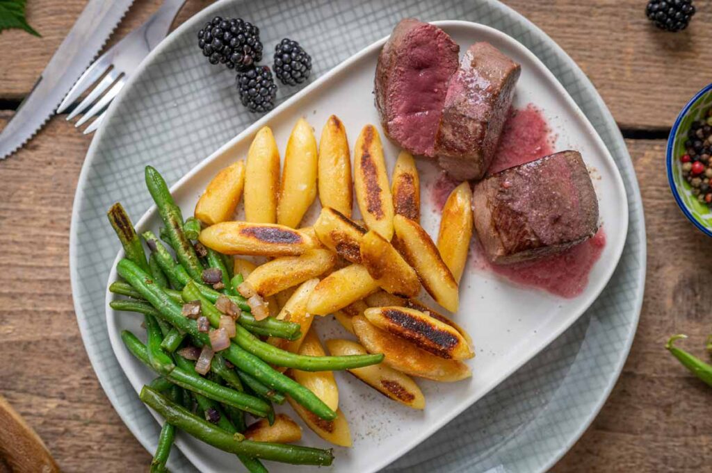 Zart rosa Rehrücken mit fruchtiger Brombeersoße, grünen Bohnen und knusprigen Schupfnudeln auf einem Teller