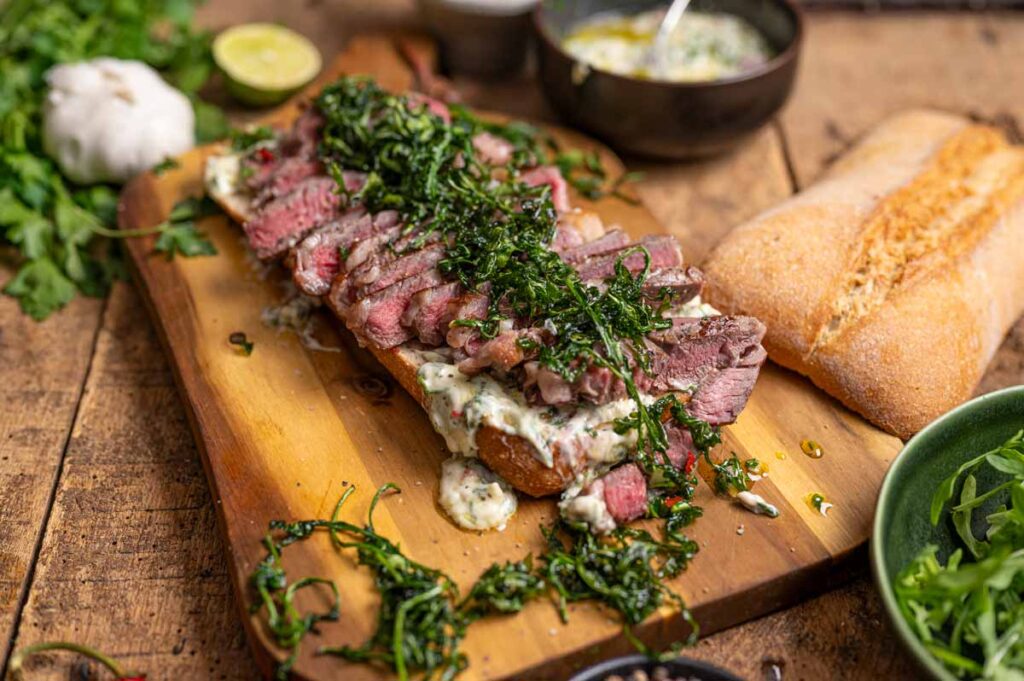 Ganzes Steak Sandwich mit goldbraunem Ciabatta, frittiertem Rucola und Chimichurri Mayonnaise auf dunklem Holzhintergrund, appetitlich präsentiert.