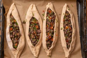 Roh geformte Pide mit Hackfleischfüllung, mit Eigelb bestrichenen Rändern und Sesam sowie Schwarzkümmel bestreut auf einem Backblech vor dem Backen